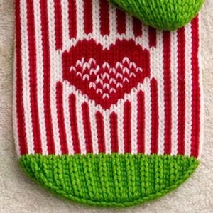 May include: Close-up of a knitted item with a red and white striped pattern. A red heart is centered on the item. The item has a green knitted cuff at the bottom. The item is likely a Christmas stocking or similar holiday decoration.