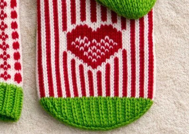 May include: Close-up of a knitted item with a red and white striped pattern. A red heart is centered on the item. The item has a green knitted cuff at the bottom. The item is likely a Christmas stocking or similar holiday decoration.