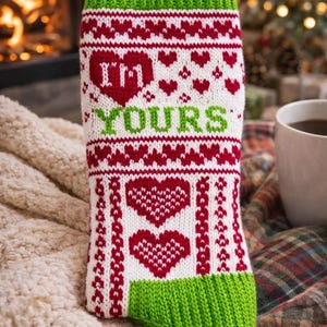 May include: A white and red knitted Christmas stocking with the words "I'M YOURS" and heart designs. The top and toe of the stocking are green. The stocking is on a cream-colored blanket.