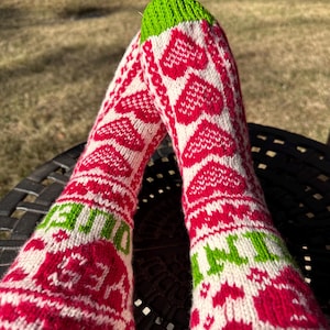 May include: A pair of knitted socks with a red and white heart pattern. The socks have green cuffs and the word "YES" is visible on the side. The socks are on a black surface.