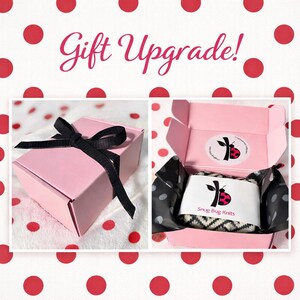 May include: Two pink gift boxes are shown, one closed with a black ribbon and the other open, revealing a wrapped item. The open box contains a black and white patterned item, and the text "Gift Upgrade!" is at the top.