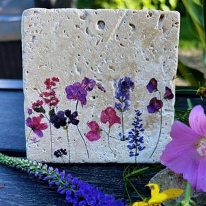 May include: A square, beige stone coaster with a floral design. The flowers are in shades of purple, pink, and red. The coaster has a rough, textured surface and is surrounded by greenery and other flowers.
