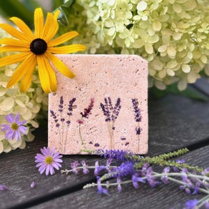May include: A square, light pink stone coaster with lavender sprig designs. The coaster is surrounded by yellow and purple flowers and green foliage, set on a dark wooden surface.