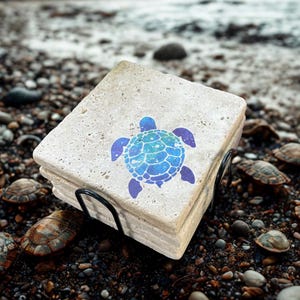 May include: A stack of square stone coasters with a blue and turquoise turtle design. The coasters are held in a black metal holder. The background is a beach scene with small rocks and pebbles.
