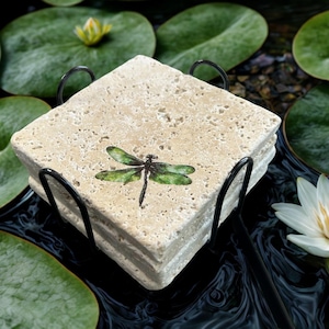 May include: A set of four square stone coasters with a black metal holder. Each coaster features a green and black dragonfly design. The coasters are beige and have a textured surface. The set is displayed near lily pads and water lilies.