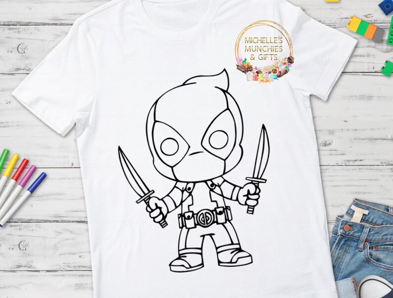 May include: A white t-shirt with a black and white outline of a cartoon superhero character holding two swords. The character is wearing a mask and a belt with a circle on it. The text "Michelle's Munchies & Gifts" is in a circle on the shirt.