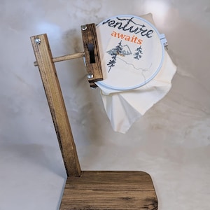 May include: Wooden embroidery hoop stand with a white hoop featuring the text "Adventure awaits" and a small mountain scene. The stand is brown and has a rectangular base.