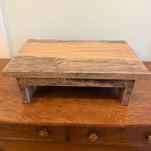 May include: A small, square wooden riser with a dark brown finish. The riser has a flat top and is supported by four short legs. The wood grain is visible, adding texture. The riser is designed to elevate items.