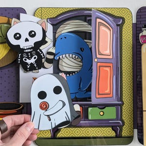 May include: A Halloween-themed children's game with a purple, green, and yellow armoire. The armoire has a door that opens to reveal a blue shark with a white bandage. A ghost with a lollipop is in front of the armoire. A skeleton cat is to the left of the armoire.