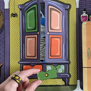 May include: A purple wooden cabinet with a green, orange, and pink door. The door is open and a blue and white mummy monster is peeking out. The cabinet has a drawer with a green card inside.