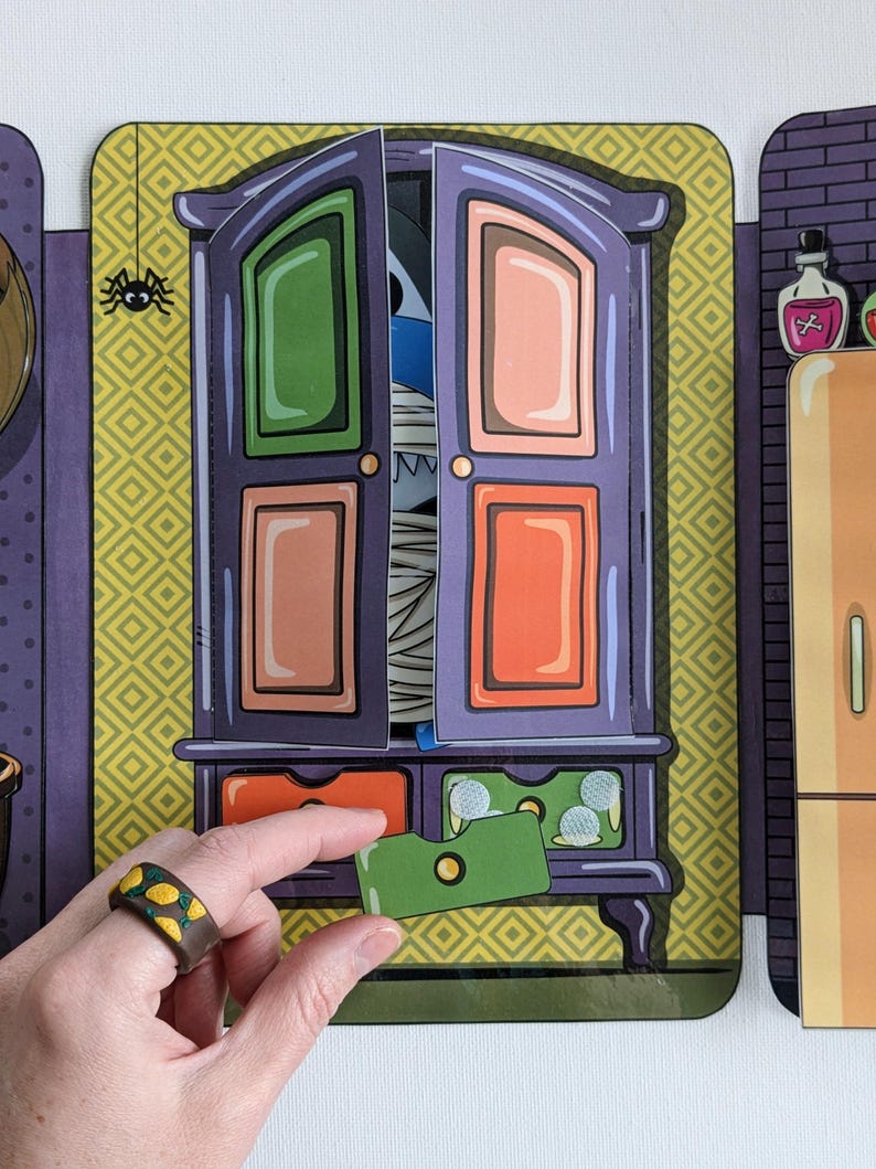 May include: A purple wooden cabinet with a green, orange, and pink door. The door is open and a blue and white mummy monster is peeking out. The cabinet has a drawer with a green card inside.