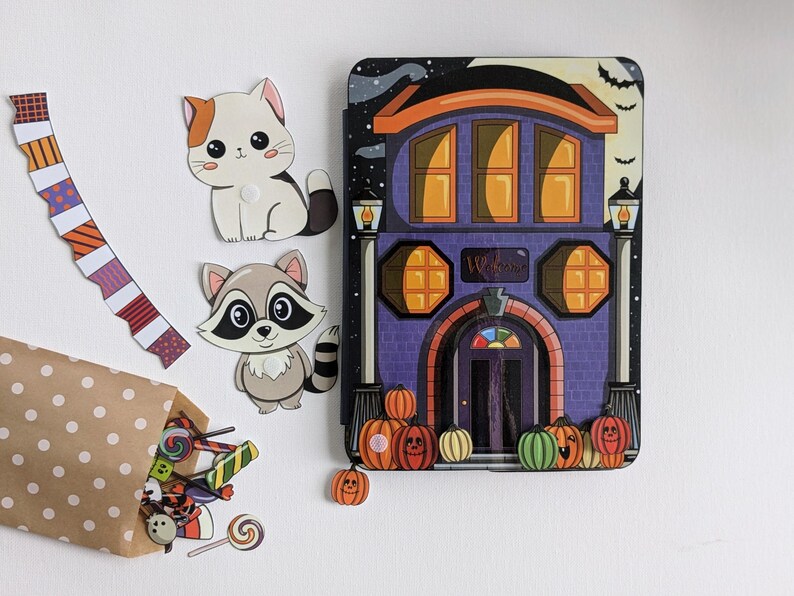 May include: A Halloween-themed playset with a purple house with orange windows and a door that says "Welcome". There are two cut-out figures of a cat and a raccoon, a brown paper bag with candy, and orange pumpkins.