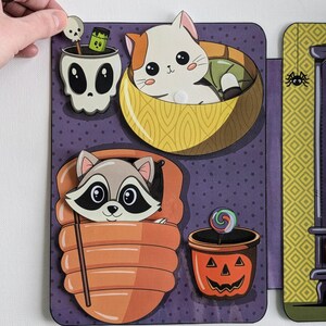 May include: A purple and yellow Halloween-themed felt board with a variety of felt cutouts. The cutouts include a raccoon in a sleeping bag, a cat in a bowl, a skull with a green potion, and a jack-o-lantern with a lollipop.