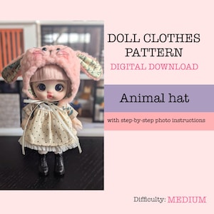 May include: A doll wearing a pink bunny hat with floral patterned ears and a cream-colored dress with black polka dots. The image includes text that reads "DOLL CLOTHES PATTERN DIGITAL DOWNLOAD Animal hat with step-by-step photo instructions Difficulty: MEDIUM."