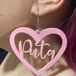 May include: Pink heart-shaped earrings with the word "Puta" written in cursive inside the heart.