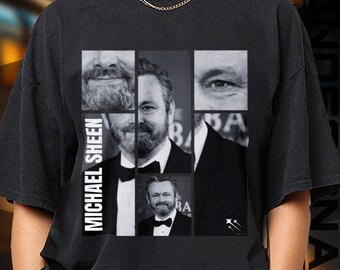 May include: Black t-shirt featuring a collage of images of Michael Sheen. The design includes close-ups of his face and a full portrait, with the name "MICHAEL SHEEN" printed vertically on the left side. The shirt is a crew neck.