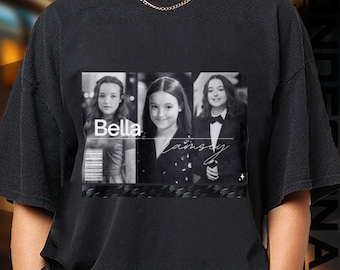 May include: Black t-shirt featuring a black and white graphic of a person in three different poses. The graphic includes the name "Bella" and a signature. The shirt is made of a soft material.