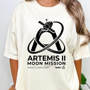 May include: Ivory-colored t-shirt featuring a black graphic of a spacecraft orbiting a planet, with the text "ARTEMIS II MOON MISSION" and "ORION LUNAR ORBIT" below. The NASA logo is also visible. The shirt is a short-sleeved, crew neck style.
