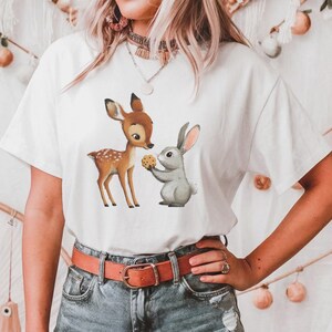 Puede incluir: Camiseta blanca con una ilustración en acuarela de un cervatillo y un conejo gris compartiendo una galleta. El cervatillo tiene manchas marrones y el conejo sostiene la galleta. La camiseta tiene mangas cortas.