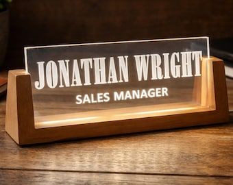 May include: A lighted desk nameplate with the name "JONATHAN WRIGHT" and the title "SALES MANAGER" in white lettering. The nameplate is made of clear acrylic and sits in a wooden base. The base is illuminated with a warm light.