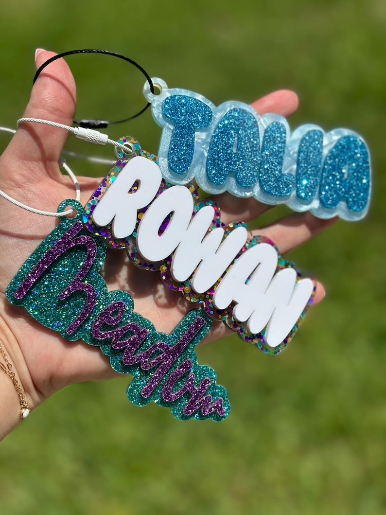 May include: Three glitter name tags in blue, teal, and purple. The names "Talia", "Rowan", and "Beachy" are displayed in a bubble font. Each tag has a loop for attaching to a keychain or bag.