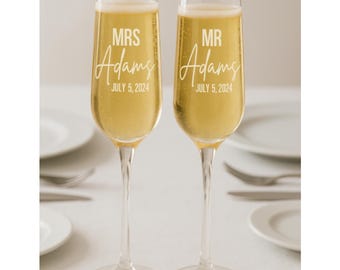 May include: Two champagne flutes filled with a golden liquid. Each glass is etched with the words "MRS Adams" and "MR Adams" along with the date "July 5, 2024". The glasses are on a white tablecloth, with plates and silverware in the background.