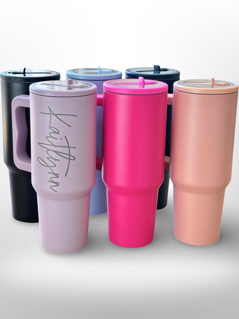 May include: An assortment of insulated tumblers in shades of black, pink, lilac, blue, and peach. Each tumbler features a handle and a lid. One tumbler is inscribed with the name "Kaitlynn".