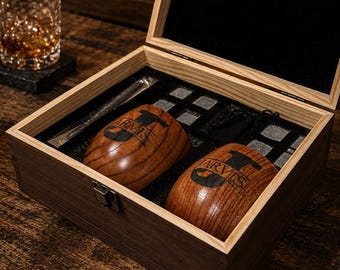 May include: A wooden gift box containing two rounded wooden cups engraved with the name JARVIS, a set of grey stone whiskey chilling cubes, and a pair of metal tongs. The interior of the box is lined with black foam inserts for organization.