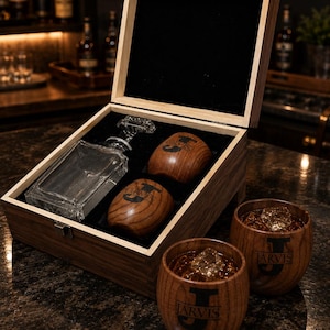 May include: A wooden presentation box with a black interior containing a clear glass decanter and two wooden cups. Two additional wooden cups filled with amber liquid and ice cubes sit outside the box. Each cup features the letter J and the name JARVIS.