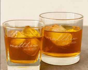 May include: Two clear whiskey glasses filled with amber liquid and ice cubes. Each glass is personalized with the name "Matthew" and the date "05.24.2025" in a delicate script. The glasses are set against a neutral background.