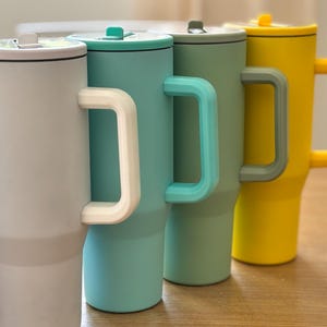May include: Four insulated travel mugs in a row, each with a handle and lid. The mugs are in shades of grey, teal, green, and yellow. The handles and lids are in coordinating colours. The mugs are designed to keep beverages hot or cold.