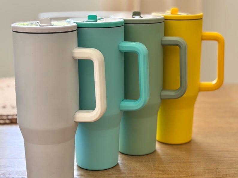 May include: Four insulated travel mugs in a row, each with a handle and lid. The mugs are in shades of grey, teal, green, and yellow. The handles and lids are in coordinating colours. The mugs are designed to keep beverages hot or cold.
