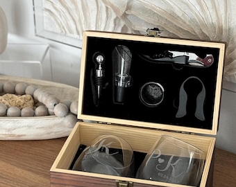 May include: A wooden wine gift set with a black interior, including a corkscrew, bottle stopper, aerator, and two engraved glasses. The glasses are etched with initials and a date. The set is presented in an open wooden box.