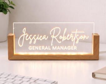 May include: A lighted desk nameplate with the name "Jessica Robertson" and the title "General Manager" in a stylish font. The nameplate is made of clear acrylic and sits in a wooden base, illuminated by warm white lights.