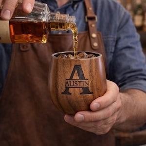 May include: A wooden tumbler with the name "Austin" and the letter "A" engraved on the front. Amber liquid is being poured into the tumbler, which contains ice cubes. The tumbler is held by a person wearing a brown apron.