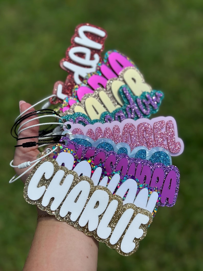May include: A hand holds a stack of glittery name tags. The tags feature names in white, gold, pink, purple, and blue glitter. Each tag has a different color scheme and is attached to a black or white cord.