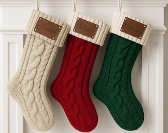 May include: Three cable-knit Christmas stockings in cream, red, and green hang from a white mantel. Each stocking has a cream-colored cuff and a brown leatherette tag with a name: Jessica, Tyler, and Madde.