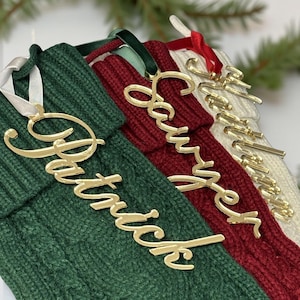 May include: Close-up of four Christmas stockings in green, red, and cream colors. Each stocking has a gold-colored name tag with names like "Patrick" and "Sawyer". The stockings are made of a knitted material.