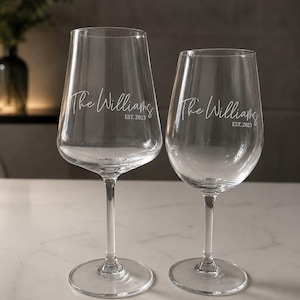 May include: Two clear glass wine glasses of different sizes, each featuring white etched text that reads The Williams and EST. 2023. The glasses have long, slender stems and wide, circular bases, standing upright with a smooth, transparent finish.