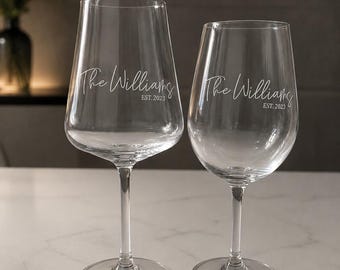 May include: Two clear glass wine glasses of different sizes, each featuring white etched text that reads The Williams and EST. 2023. The glasses have long, slender stems and wide, circular bases, standing upright with a smooth, transparent finish.
