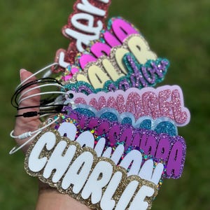 May include: A stack of colorful glitter name tags with white lettering. The names on the tags are "Audrey", "Olivia", "Sophia", "Summer", "Emma", and "Charlie".