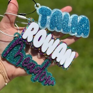 May include: Three personalized glitter keychains with the names "Talia", "Rowan", and "Reagan" in white letters. The keychains are made of clear resin with glitter embedded in the resin. The keychains are attached to silver metal rings.
