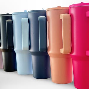 May include: A selection of insulated tumblers in shades of pink, black, light blue, navy, peach, and hot pink. Each tumbler features a matching handle and lid, suitable for drinks. The tumblers are approximately 23 cm tall.