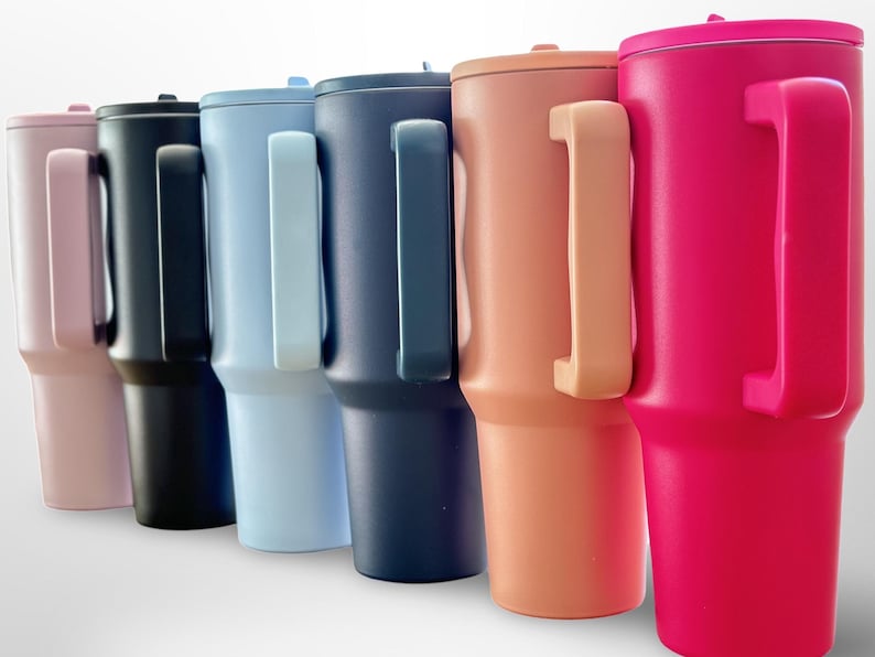May include: A selection of insulated tumblers in shades of pink, black, light blue, navy, peach, and hot pink. Each tumbler features a matching handle and lid, suitable for drinks. The tumblers are approximately 23 cm tall.