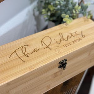 May include: A rectangular bamboo box with a metal clasp. The box is engraved with the words "The Riders" and "EST. 2024" in a cursive font. The box is on a dark wooden surface.