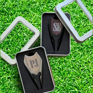 May include: Two silver golf ball divot tools in black boxes with personalized initials. The initials are "PJ" and "SE".