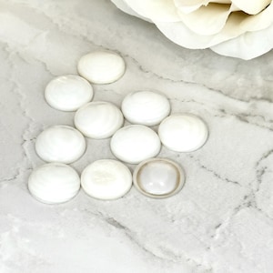 May include: A group of ten white round iridescent shell cabochons, with one cabochon in the centre that is slightly larger than the others.