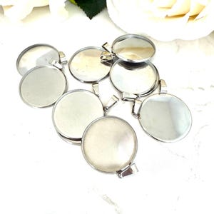 May include: A group of eight silver round pendant trays with a smooth, shiny finish. The trays are designed to hold a variety of materials, such as resin, glass, or other decorative elements.