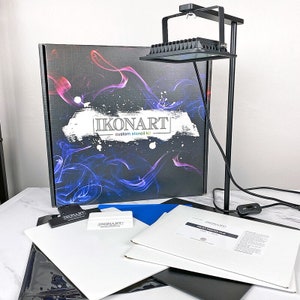 May include: A black light box with a stand and a power cord. The light box is on a white surface. There are several white sheets of paper and a blue sheet of paper on the surface. The box has the text "IKONART custom stencil kit" on it.
