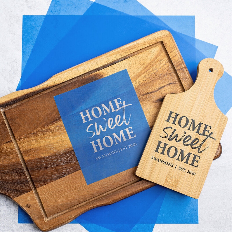 May include: Two wooden chopping boards with the text "Home Sweet Home" and the name "Swansons | Est. 2020" on them. The boards are on a blue background.